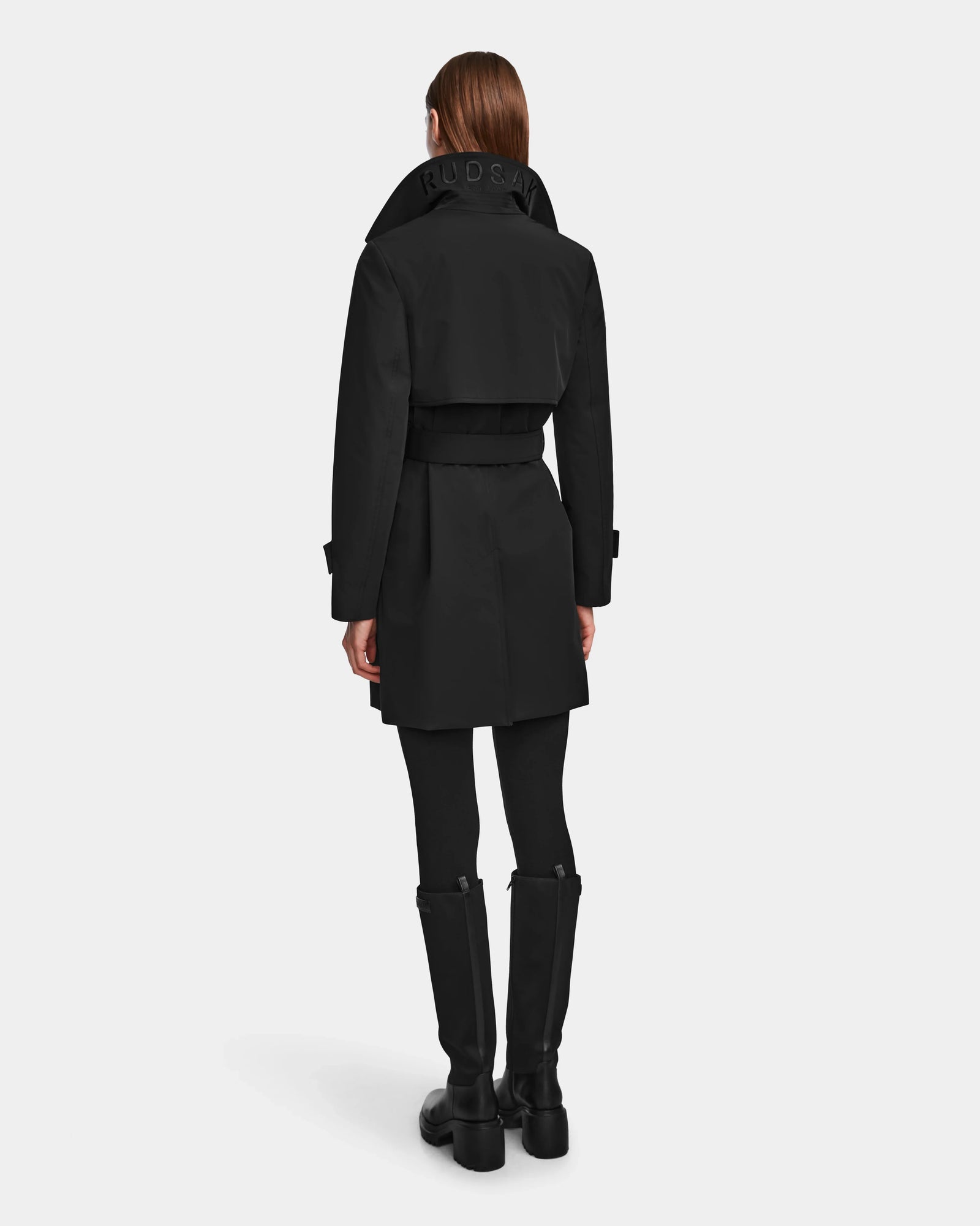A person with straight brown hair wearing the Rudsak KIMBERLY - 8124801 Black womens belted trench coat, black leggings, and knee-high black boots stands facing away from the camera against a plain light background.