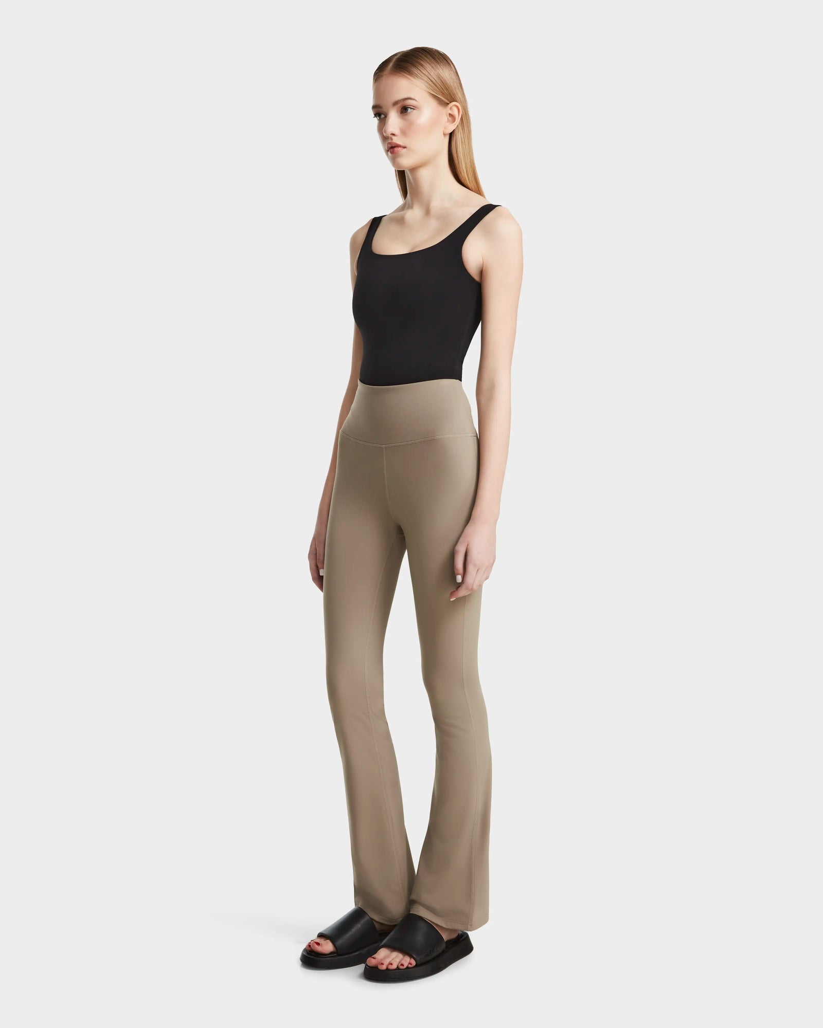 A woman with light skin and long blonde hair poses against a plain background wearing Rudsak’s ADRIANA - 8524009 Cement high-waisted beige flared-leg leggings, paired with a black tank top and black slide sandals.