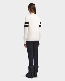 Seen from behind, a person with long straight hair wears Rudsaks ODESSA - 8523540 Iceland soft-knit après-ski sweater with two black sleeve stripes, black pants, and black winter boots against a light gray background.