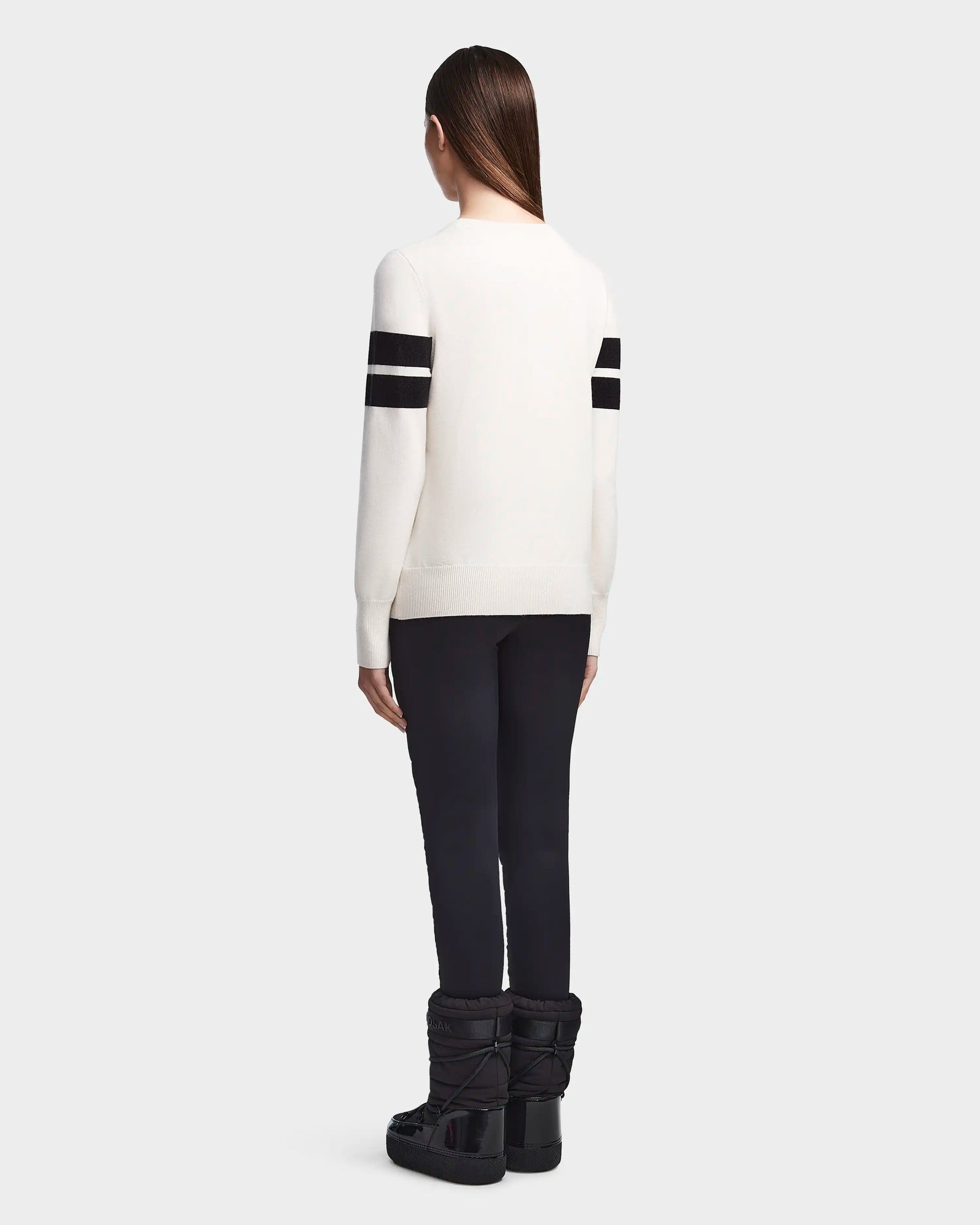 Seen from behind, a person with long straight hair wears Rudsaks ODESSA - 8523540 Iceland soft-knit après-ski sweater with two black sleeve stripes, black pants, and black winter boots against a light gray background.