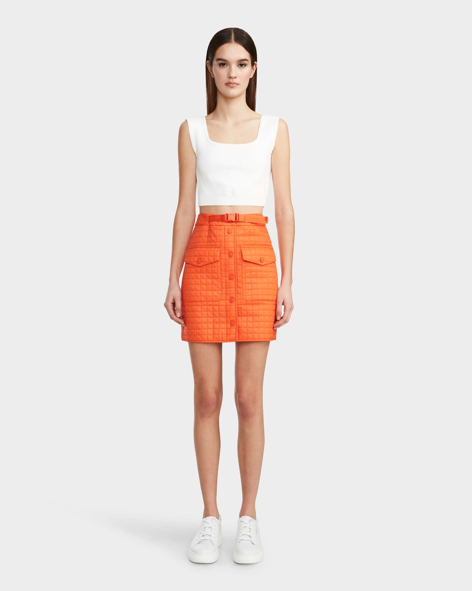 A woman faces forward in a Rudsak SABINE - 8524063 Orange Sorbet quilted mini skirt with buttons, pockets, and a matching belt, paired with a sleeveless white top and white sneakers against a light gray background.