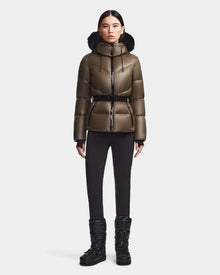 A woman faces forward wearing the Rudsak CHRISTELLE S - L123524 Light Olive belted puffer jacket with a fur-trimmed hood, paired with black leggings and winter boots, set against a plain light background.