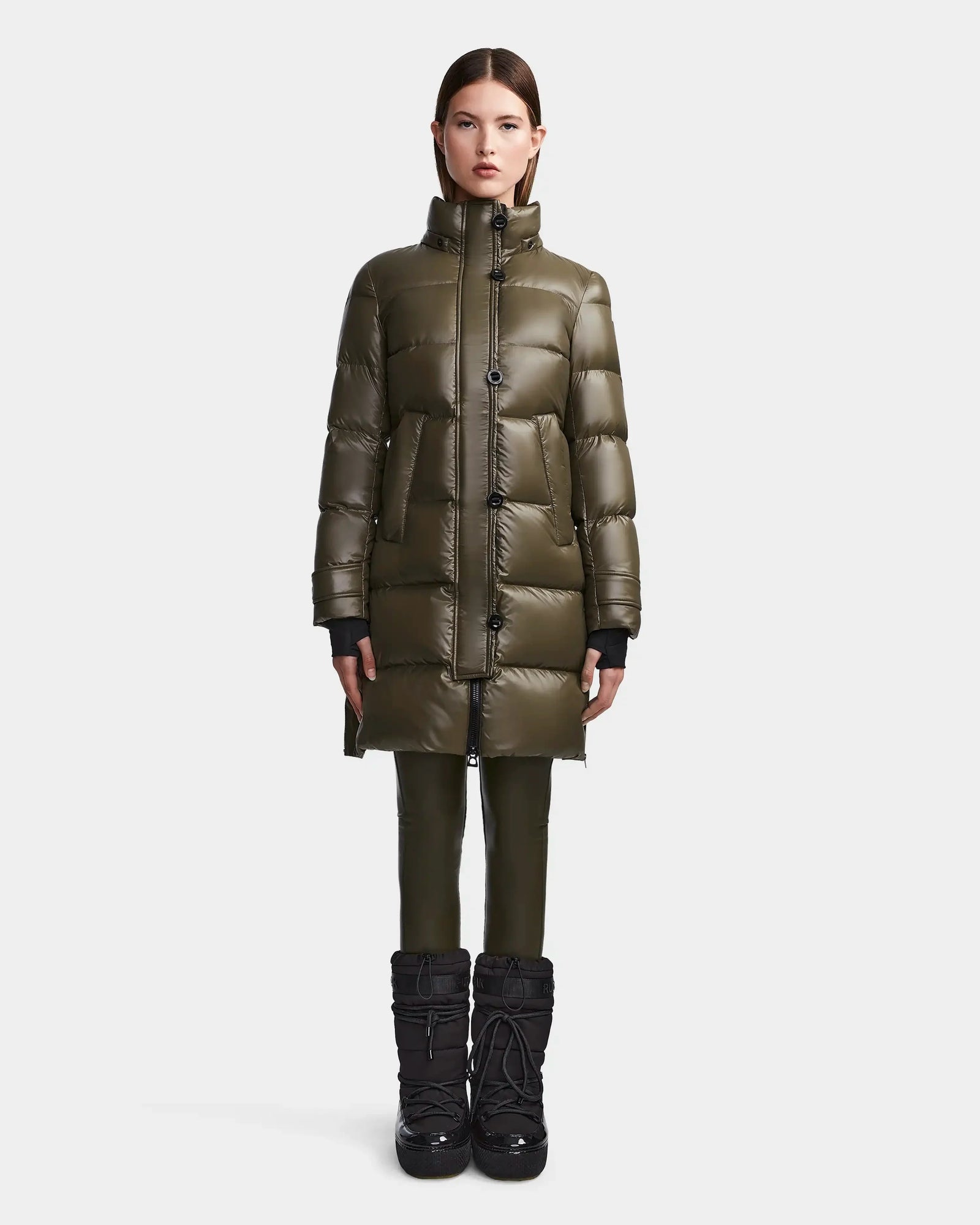 A woman faces forward against a plain white background, wearing the Rudsak MEYA S - L123521 Light Olive women's down puffer with large pockets, paired with black pants and black winter boots.