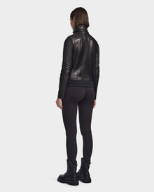 A woman with chin-length brown hair wears the Rudsak PRESLEY - 8122027 BLACK leather jacket, paired with black leggings and boots, standing with her back to a plain light background.