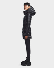A person stands in profile against a plain light background, wearing the Rudsak MALAYA - L123510 Black women’s long, hooded down puffer jacket with herringbone quilting, paired with black leggings and chunky boots.