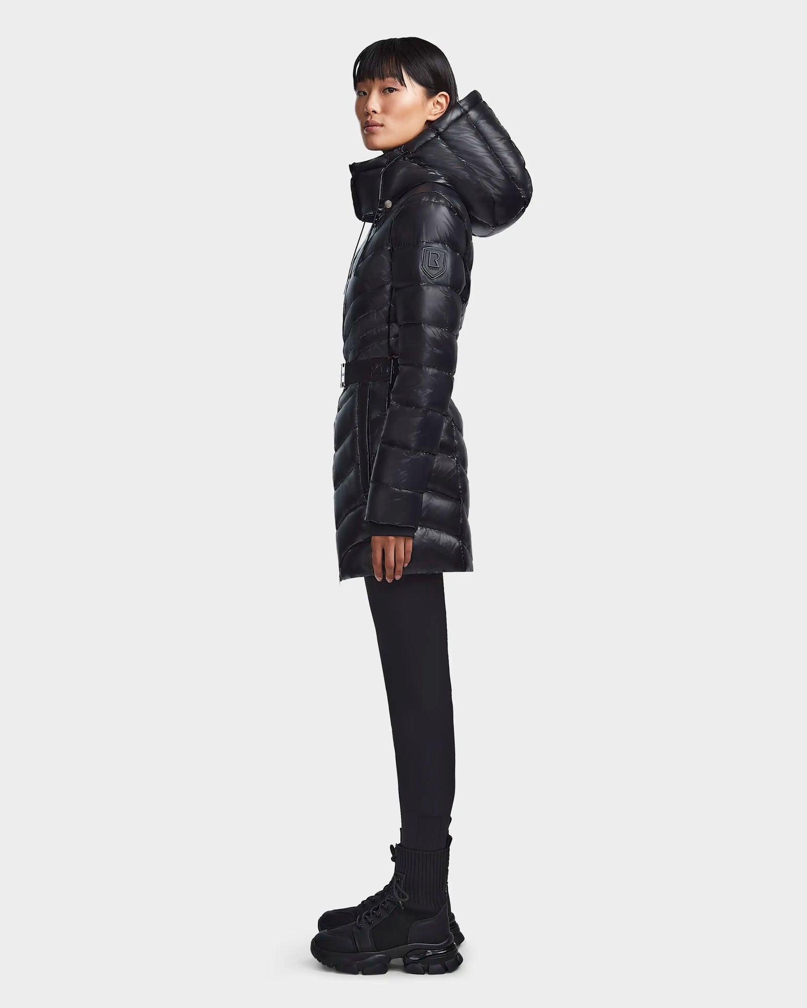 A person stands in profile against a plain light background, wearing the Rudsak MALAYA - L123510 Black women’s long, hooded down puffer jacket with herringbone quilting, paired with black leggings and chunky boots.
