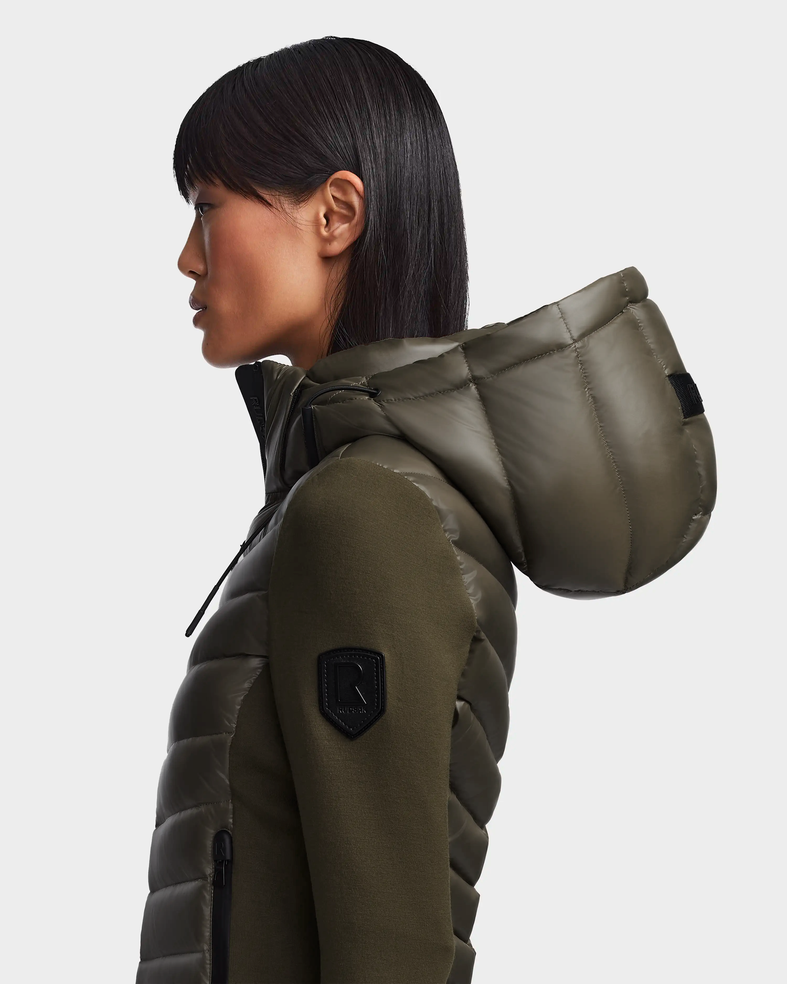 A woman with straight dark hair, seen in profile against a plain light background, wears the Rudsak MALYA - L121006 Light Olive activewear-inspired quilted hooded jacket featuring a black sleeve patch.