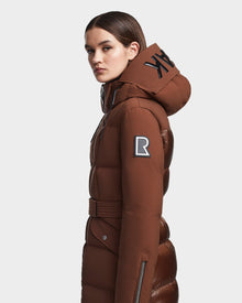 A woman in a brown CALI - 8122502 COHIBA down puffer coat by RUDSAK, belted with a hood, large black letters, and a black-and-white R patch on the sleeve, stands sideways against a plain background.
