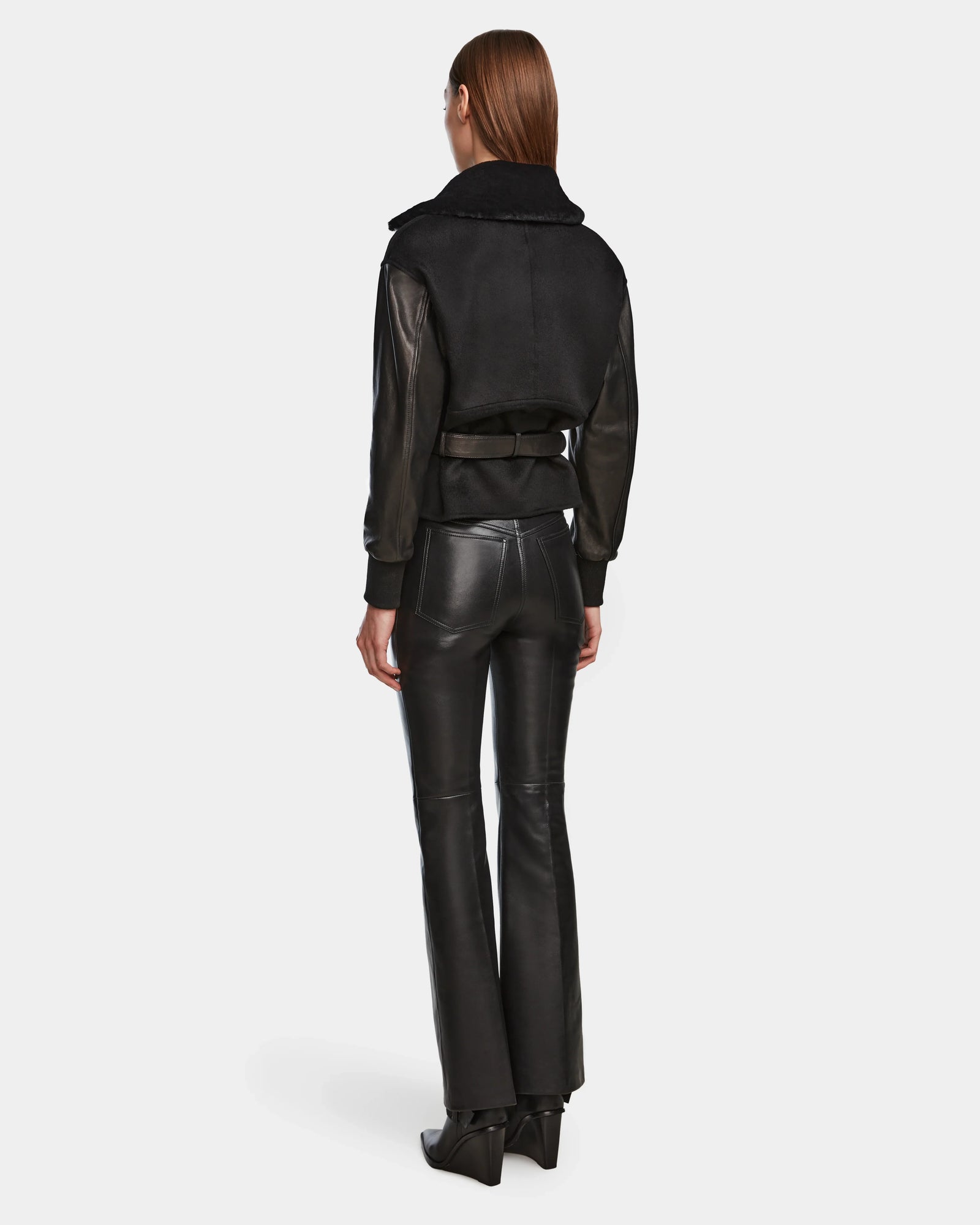 A woman with straight brown hair faces away, wearing the Rudsak HALA - 8124521 Black women’s shearling jacket with a wide collar and belt detail, paired with black leather pants and pointed-toe boots against a plain light background.