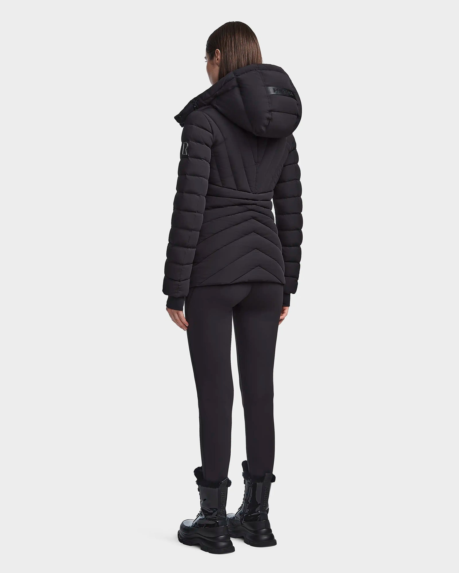 A person wearing the Rudsak HANNA - 8123562 Black winter down jacket stands facing away from the camera, paired with black leggings and boots, against a plain white background.