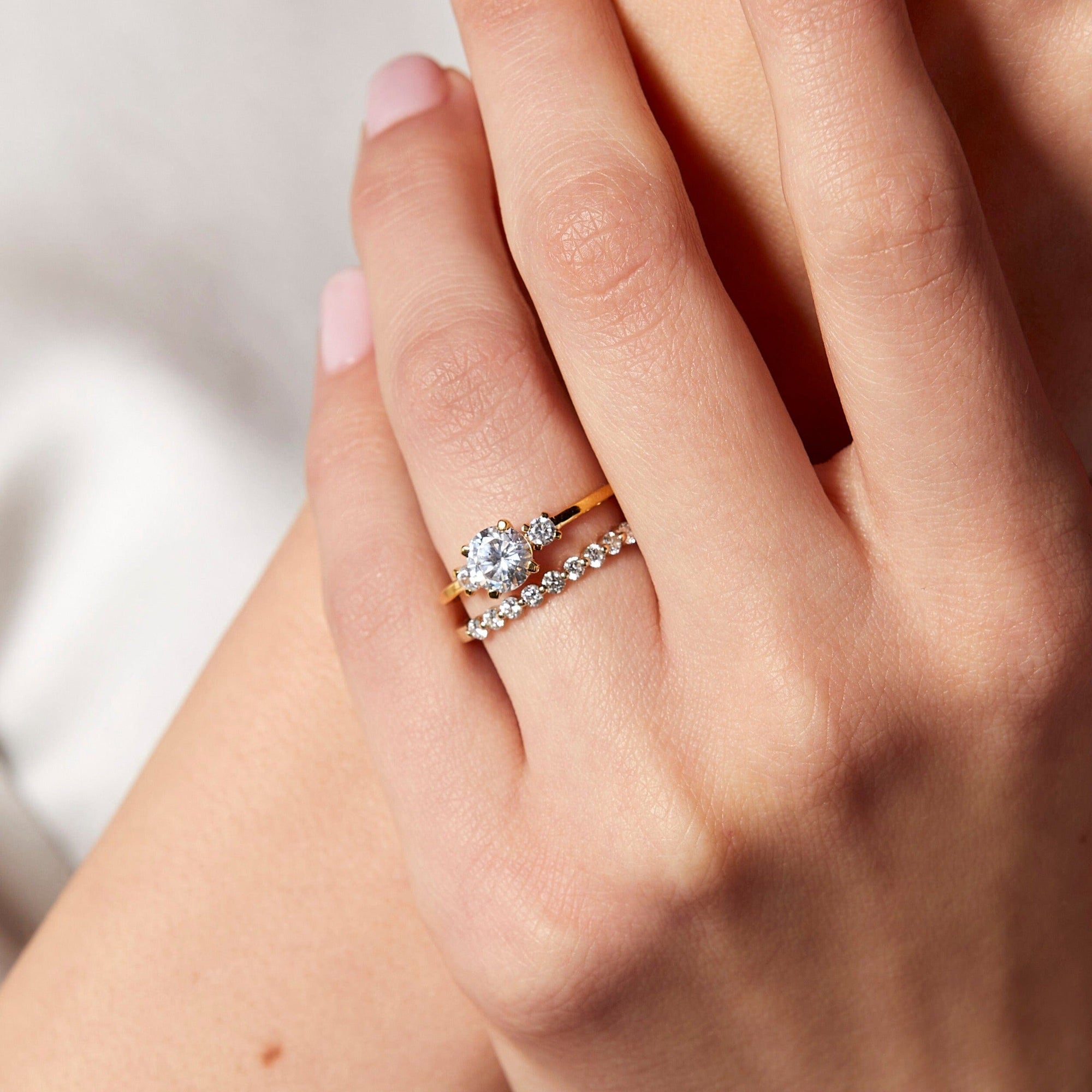 Lifestyle Model Close-up of a woman's hand resting gracefully against a soft white fabric. On her ring finger, she is wearing a stack of two gold rings: a three-stone diamond ring featuring a larger round center diamond flanked by two smaller diamonds, paired with a delicate prong-set diamond eternity band below it.